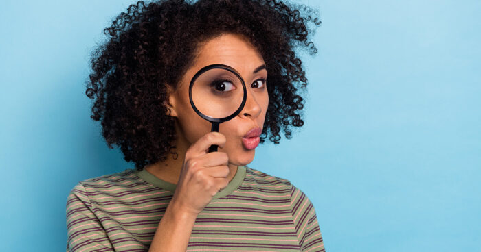 woman in a striped shirt holding a magnifying glass to her face with a curious expression as she looks through it, one eye magnified through the lens.