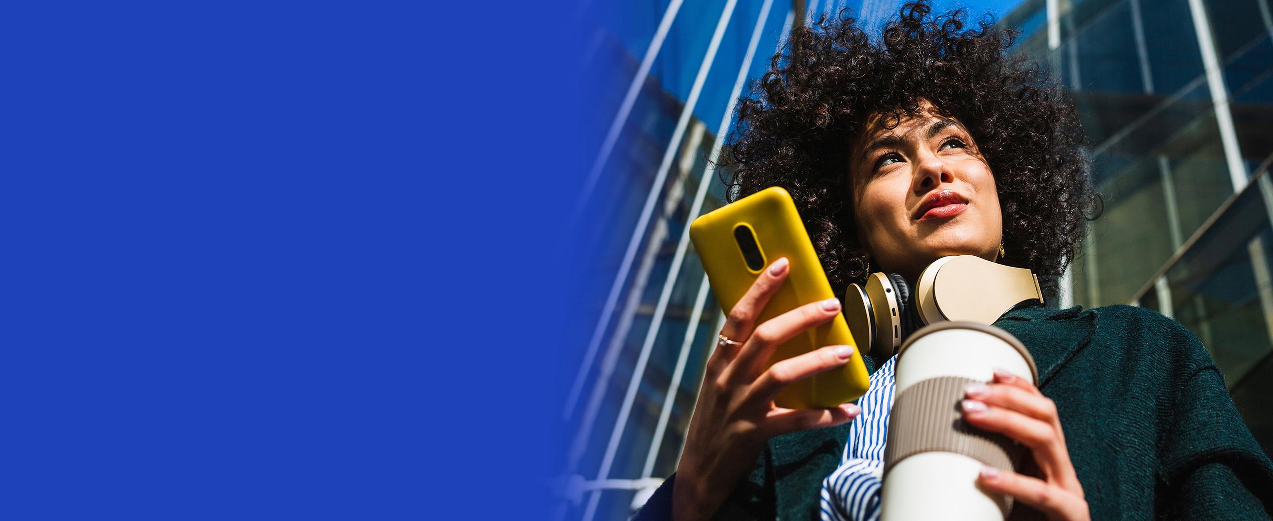 Low angle view of businesswoman looking to the side while texting on her cell phone, office buildings in the background. Smart woman in casual clothes against blue sky in financial district.