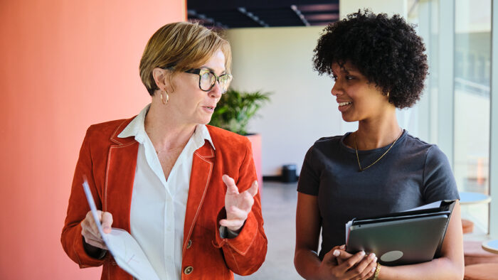 Two businesswomen of different ages smiling and talking together while walking down a corridor