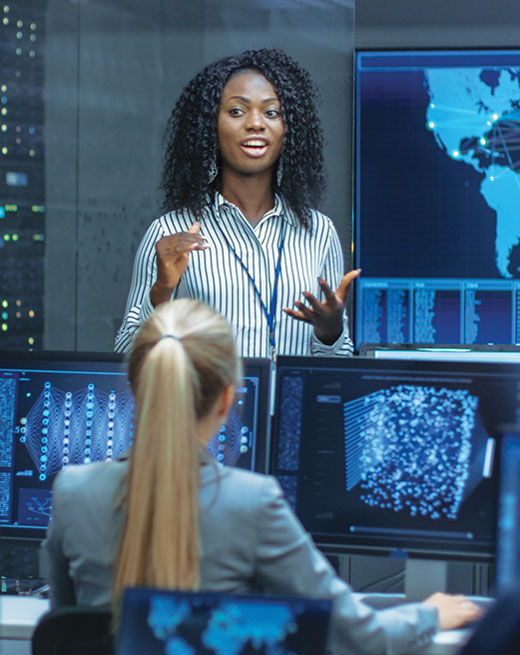 woman standing and speaking in front of a room of people seated at computer monitors