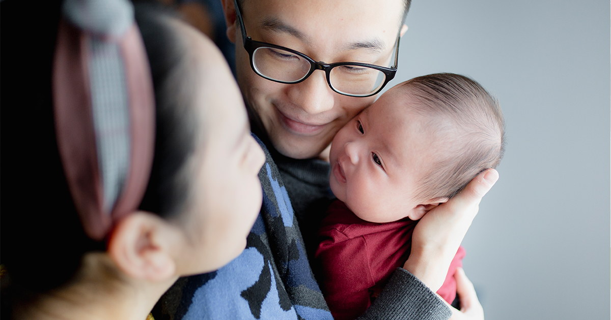 A man holds his young baby while the mother leans over his shoulder.